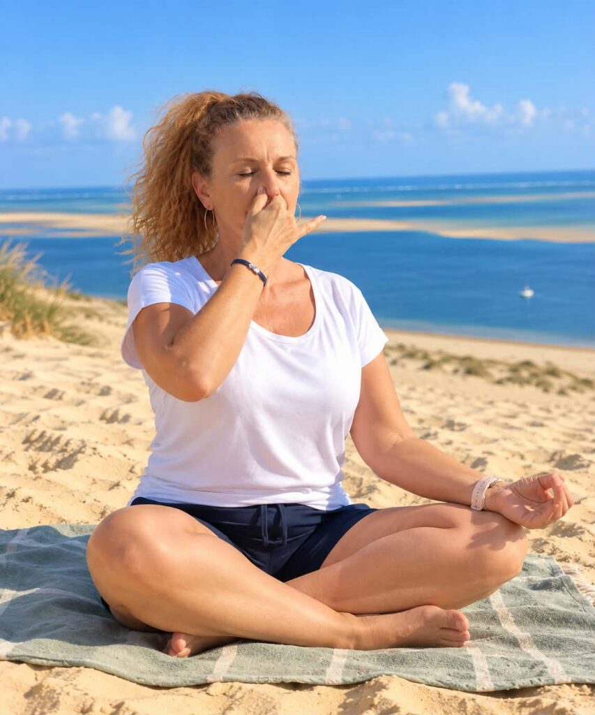 Femme méditant sur la plage, pratiquant une technique de respiration, vêtue d'un t-shirt blanc et d'un short noir, assise sur une serviette.