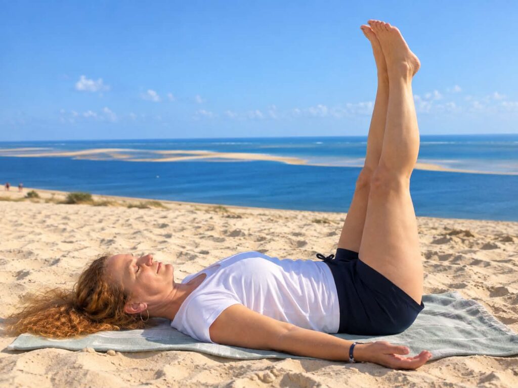 Femme allongée sur le dos sur une serviette sur la plage, jambes levées vers le ciel, vue sur la mer.