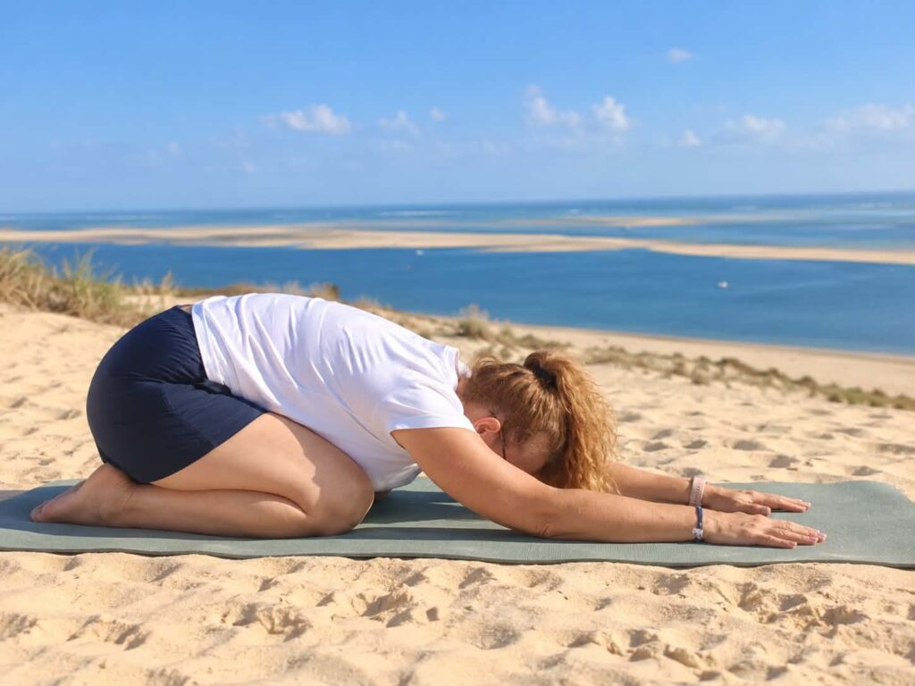 Femme pratiquant le yoga en position de l'enfant sur un tapis sur la plage, avec vue sur la mer.