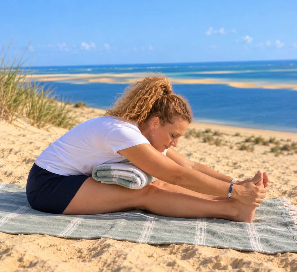 Femme en train de s'étirer sur une serviette au bord de l'eau, avec un paysage de dunes et de mer en arrière-plan.